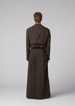 A person faces away wearing IM MEN’s SHRINK WOOL: a long, dark brown crepe-look coat layered with a cropped, collarless jacket, set against a plain light gray background.