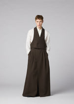 A person stands against a plain background, wearing IM MEN’s SHRINK WOOL: a white shirt with a brown crepe-look vest and wide, floor-length brown trousers, hands in pockets and a neutral expression.