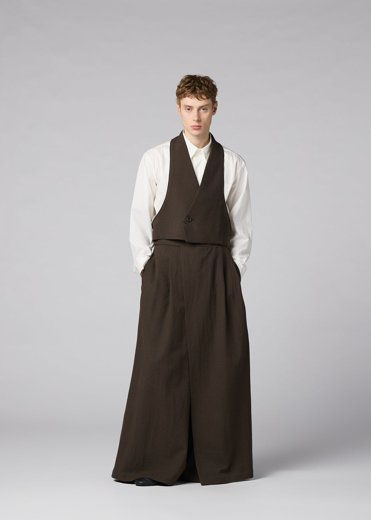 Zoomed view A person stands against a plain background, wearing IM MEN’s SHRINK WOOL: a white shirt with a brown crepe-look vest and wide, floor-length brown trousers, hands in pockets and a neutral expression.