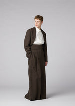 A person models the IM MEN SHRINK WOOL dark brown crepe-look long skirt suit with a white dress shirt, standing against a light gray background and looking to the side with one hand in their pocket.