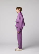 A person stands in profile against a plain background, wearing the IM MEN FRONT BACK purple suit, featuring a loose-fitting, cape-like wool jacket and matching pants. They have short hair and white shoes, demonstrating the ensemble’s versatile styling for any occasion.