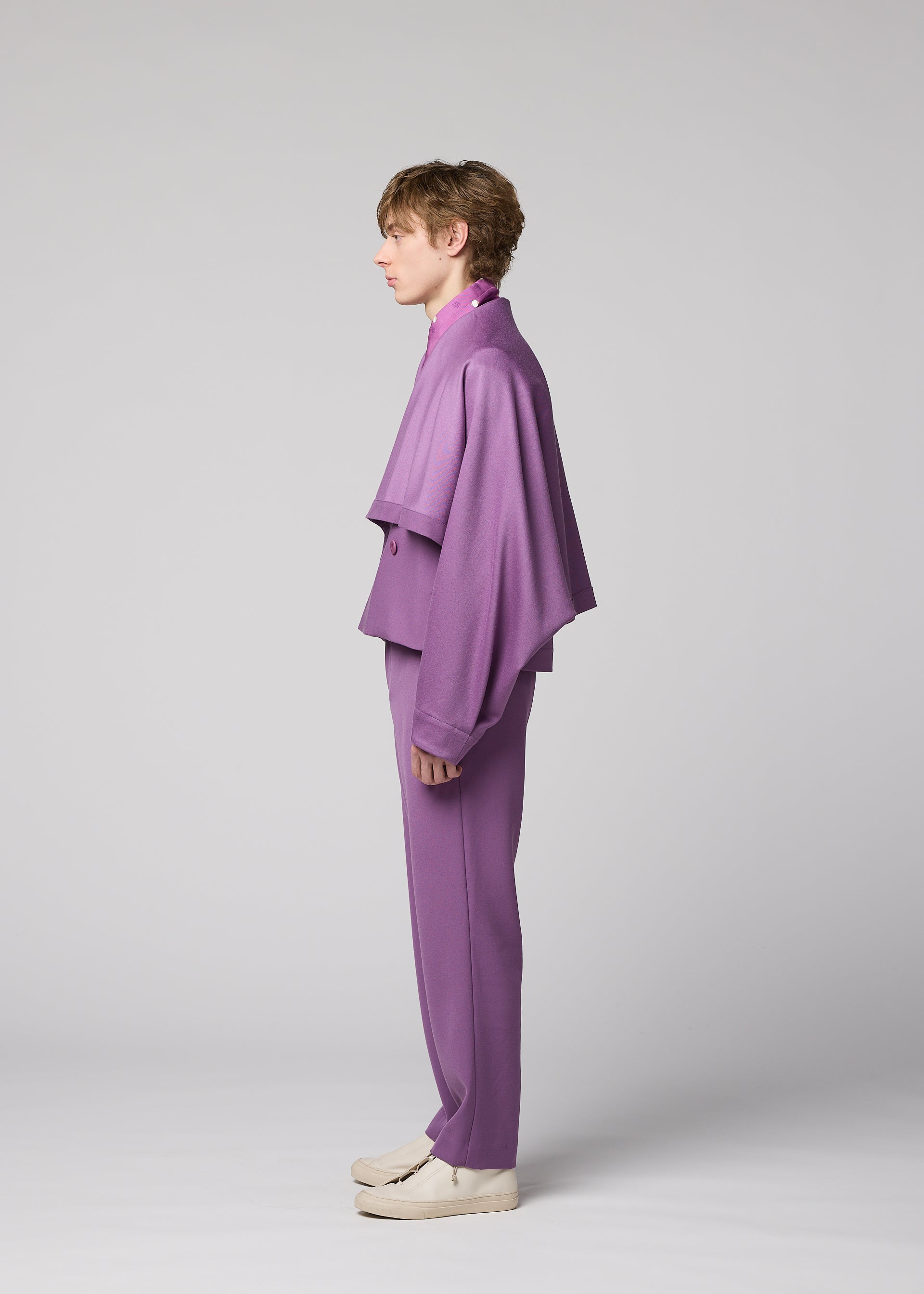 Zoomed view A person stands in profile against a plain background, wearing the IM MEN FRONT BACK purple suit, featuring a loose-fitting, cape-like wool jacket and matching pants. They have short hair and white shoes, demonstrating the ensemble’s versatile styling for any occasion.