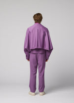 A person stands facing away against a plain gray background, wearing the IM MEN “FRONT BACK” ensemble: a loose-fitting lavender suit with a cape-like top and matching pants crafted from satin back fabric for a subtle sheen, paired with white shoes.