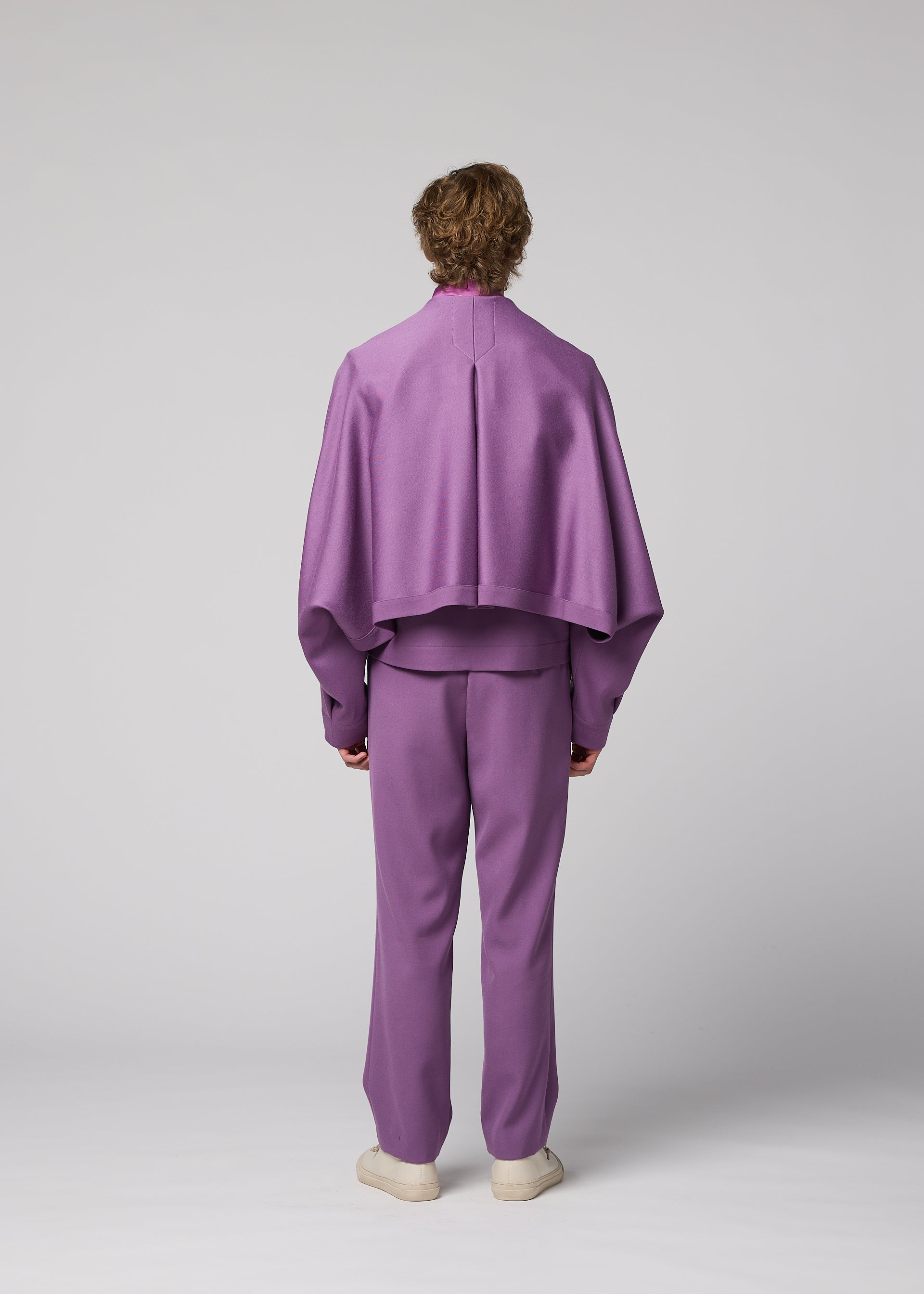 Zoomed view A person stands facing away against a plain gray background, wearing the IM MEN “FRONT BACK” ensemble: a loose-fitting lavender suit with a cape-like top and matching pants crafted from satin back fabric for a subtle sheen, paired with white shoes.