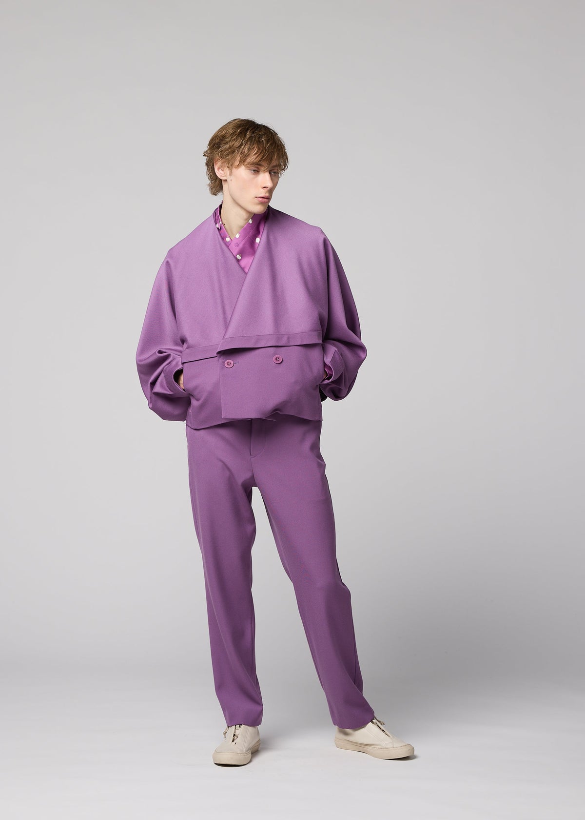 A person stands in a studio against a plain gray background, showcasing versatile styling in the IM MEN “FRONT BACK” set: a loose-fitting, double-breasted purple wool jacket paired with matching pants and completed with cream-colored shoes. view 7 of 9