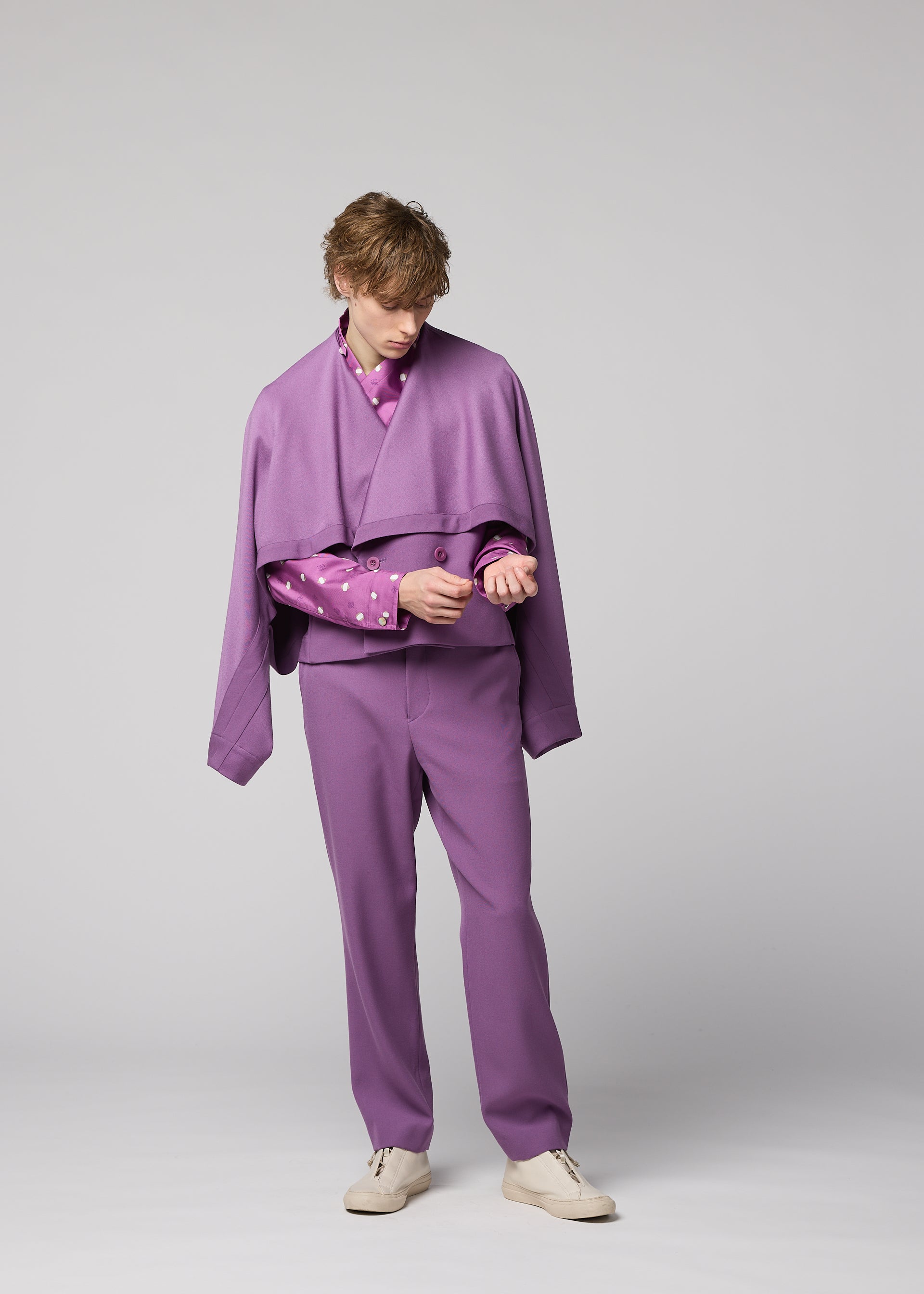 Zoomed view A person stands wearing IM MEN’s FRONT BACK purple wool jacket suit with a matching cape draped over their shoulders and crisp white shoes. As they look down, they adjust the sleeve of a pink shirt with yellow dots peeking from beneath the suit. The plain, light gray background highlights the versatile styling of this standout ensemble.