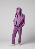 A person stands against a plain gray background wearing the IM MEN FRONT BACK look: purple pants and a matching purple polka-dot shirt, paired with a coordinating wool jacket featuring a satin back, draped over their head. One hand is in their pocket and they complete the outfit with beige shoes.