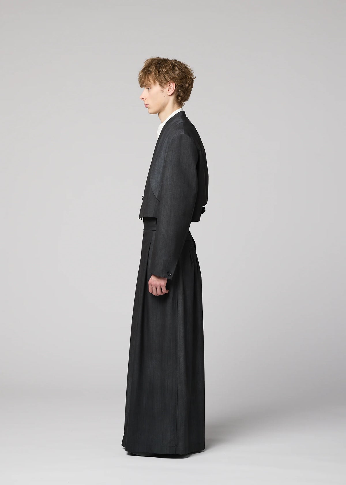 A person stands in profile against a plain background, wearing the IM MEN GRADATION WOOL tailored suit with a cropped jacket and wide, floor-length trousers from the Autumn Winter 2026/27 collection. view 10 of 13