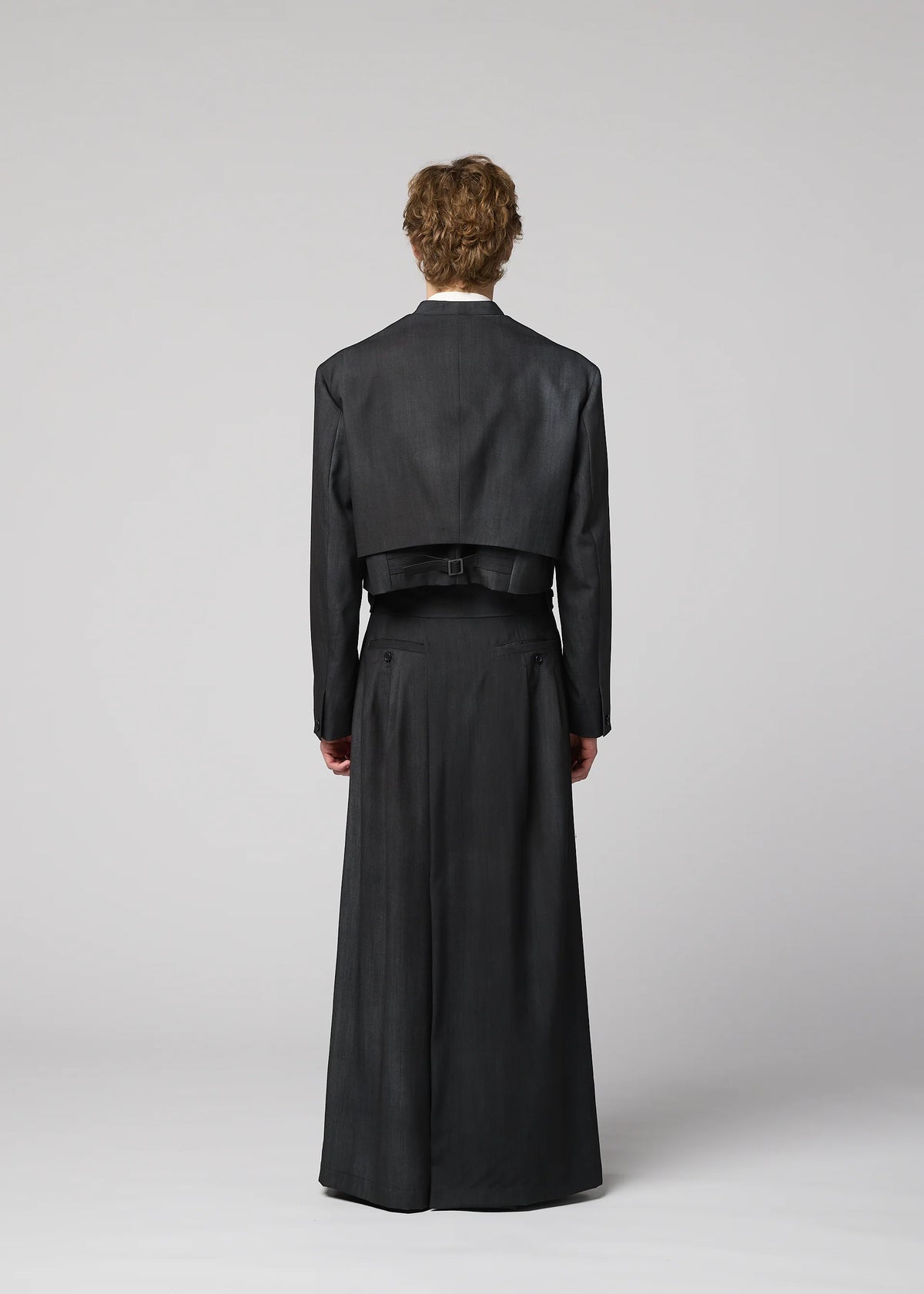 A person with short curly hair faces away, wearing IM MEN’s GRADATION WOOL dark cropped jacket and long matching skirt. The background is plain, light gray. view 11 of 13