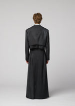 A person with short curly hair faces away, wearing IM MEN’s GRADATION WOOL dark cropped jacket and long matching skirt. The background is plain, light gray.