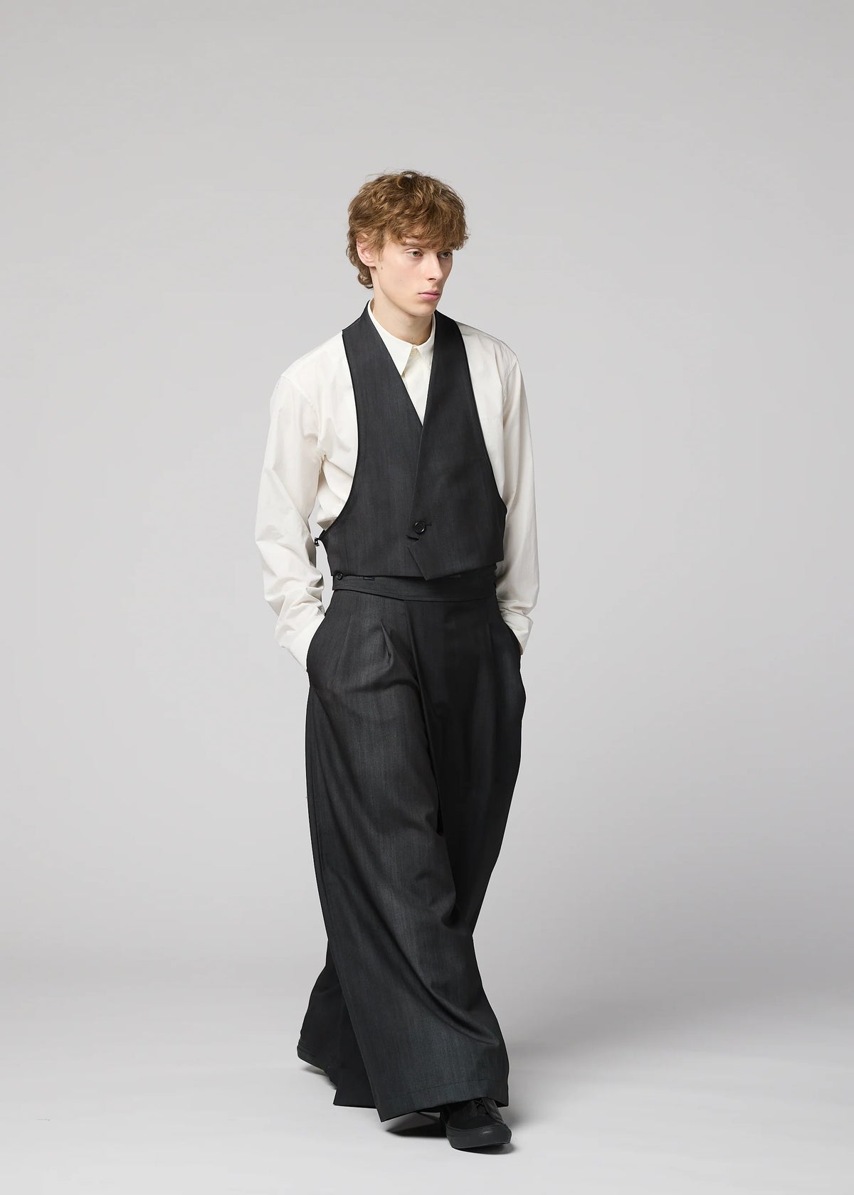 A person with wavy hair in a loose white shirt, unique vest, and dark wide-legged suit—featuring GRADATION WOOL by IM MEN—stands against a plain light gray background, hands in pockets. view 12 of 13
