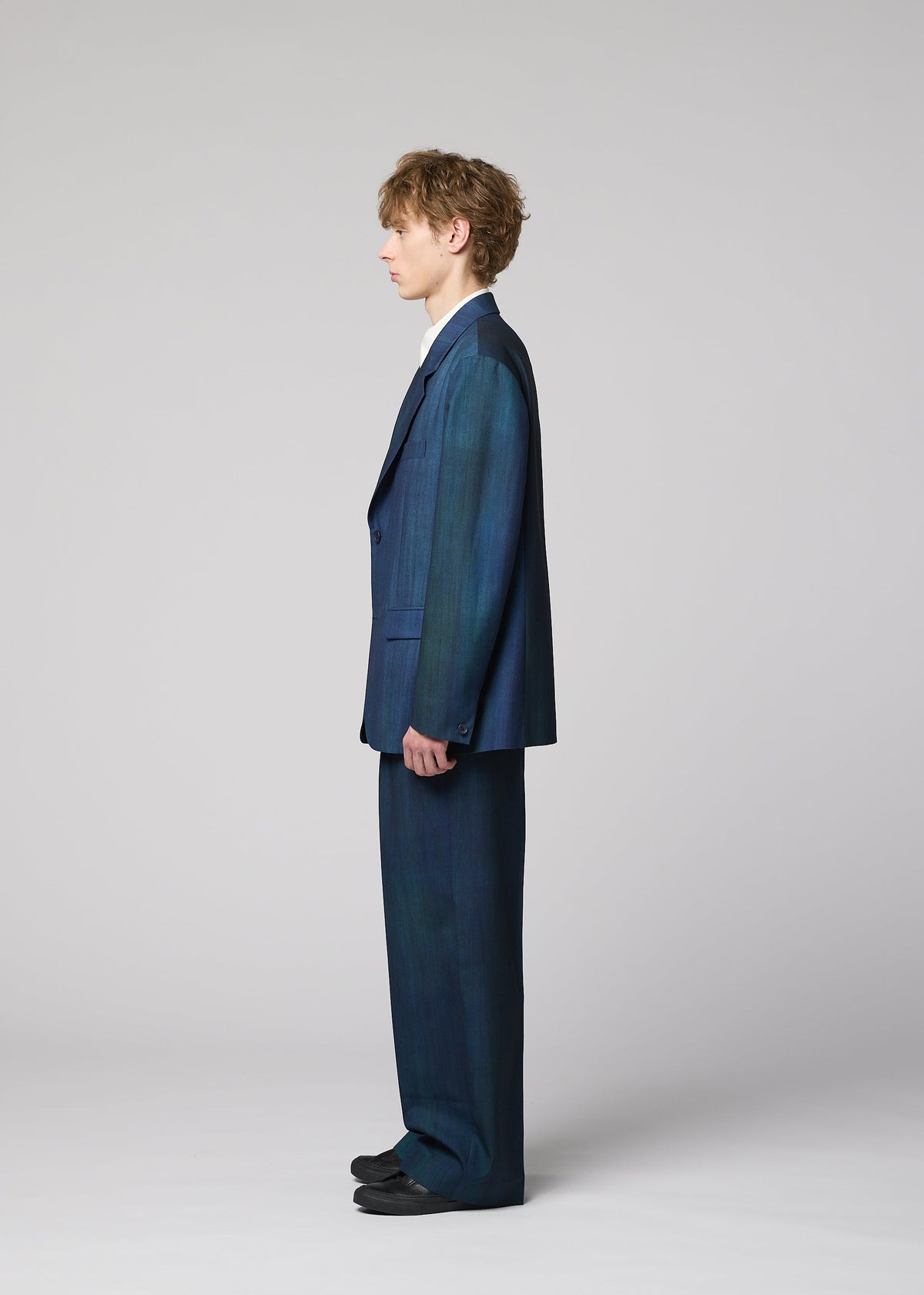 A person with wavy hair stands in profile wearing the IM MEN GRADATION WOOL jacket and blue suit from the Autumn Winter 2026/27 collection, paired with a white shirt and black shoes against a light gray background. view 6 of 8
