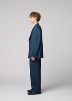 A person with wavy hair stands in profile wearing the IM MEN GRADATION WOOL jacket and blue suit from the Autumn Winter 2026/27 collection, paired with a white shirt and black shoes against a light gray background.