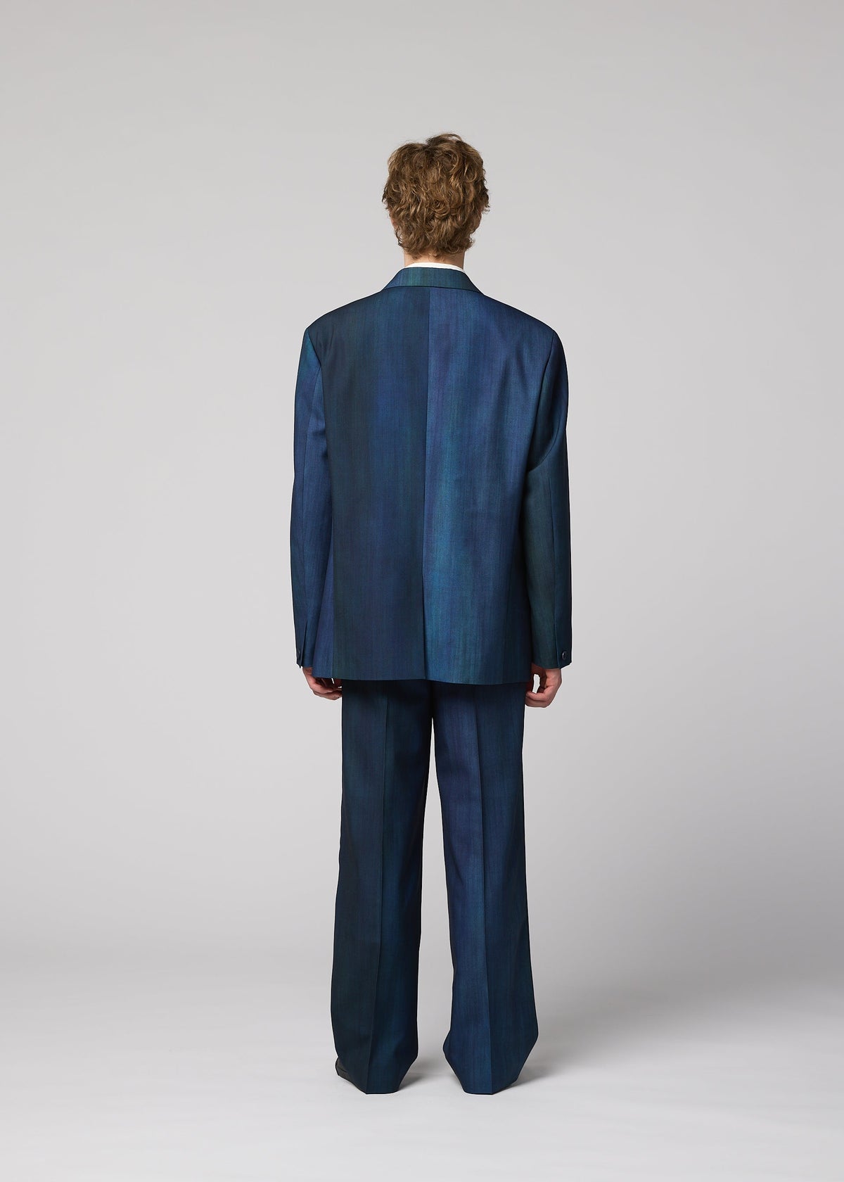 Zoomed view A person with short, curly hair faces away, wearing the IM MEN GRADATION WOOL jacket and wide-leg pants in dark blue against a plain light gray background.