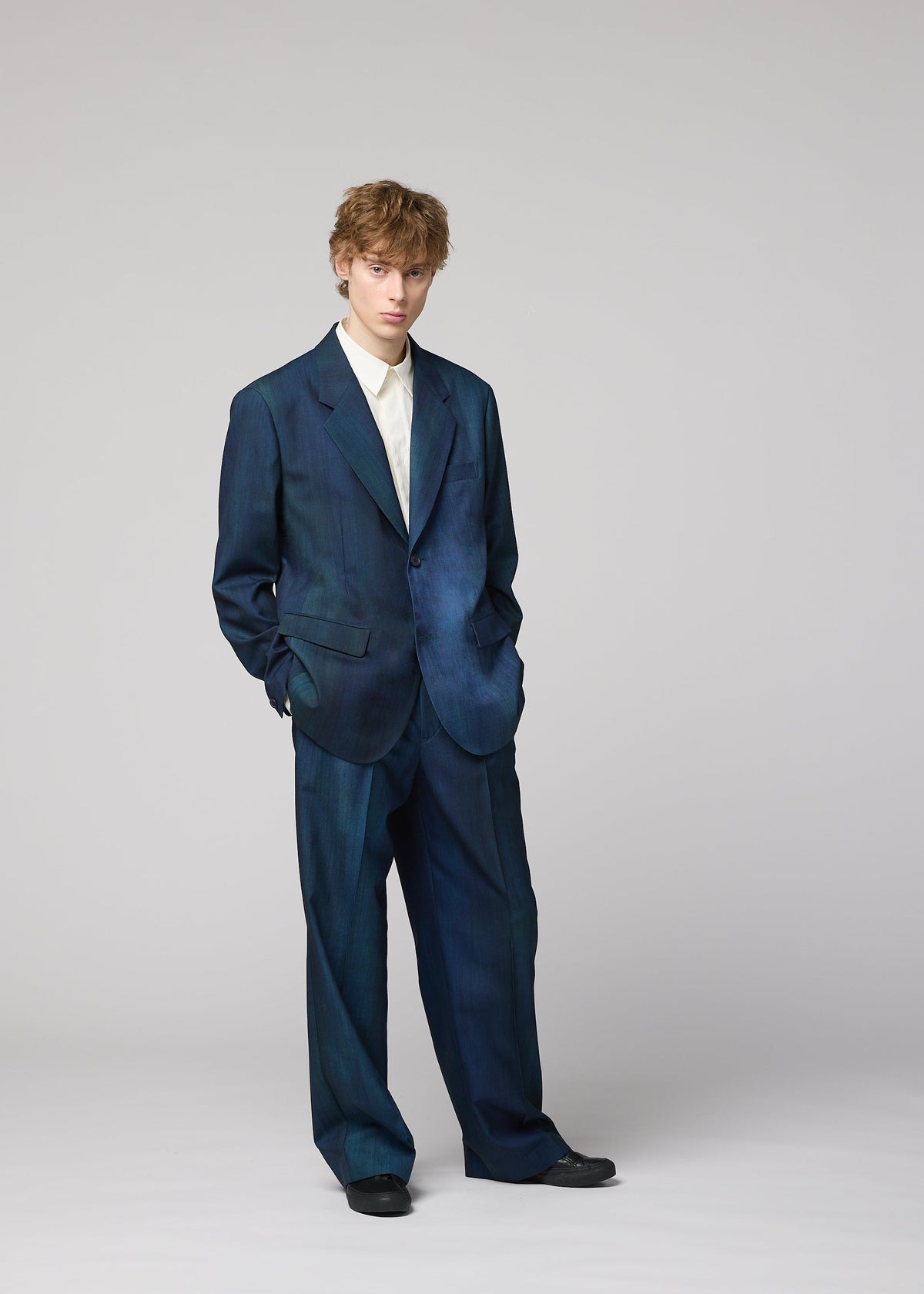 Zoomed view A person with light brown hair wears the oversized, dark blue GRADATION WOOL jacket by IM MEN, hands in pockets, looking at the camera.