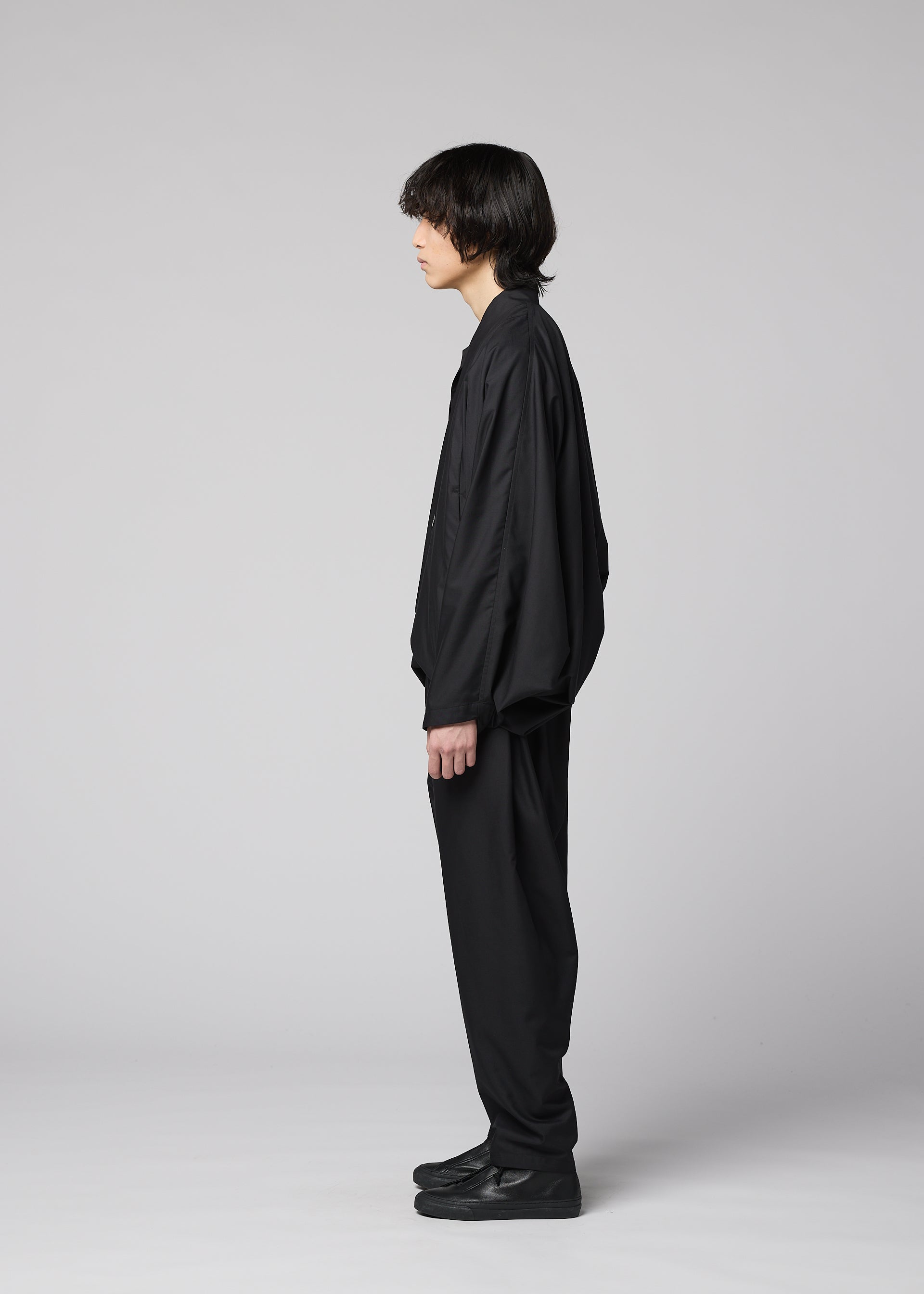 Zoomed view A person with medium-length dark hair stands in profile against a gray background, wearing IM MEN’s SWING BLACK wrinkle-resistant lightweight pants, a loose black shirt, and black shoes.