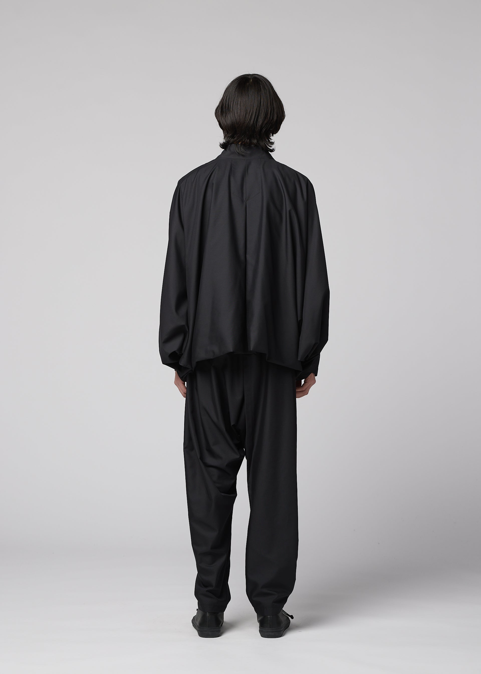 Zoomed view A person with medium-length dark hair faces away, wearing IM MEN's SWING BLACK: a loose black outfit paired with wrinkle-resistant, lightweight pants and black shoes, all set against a plain light gray backdrop.