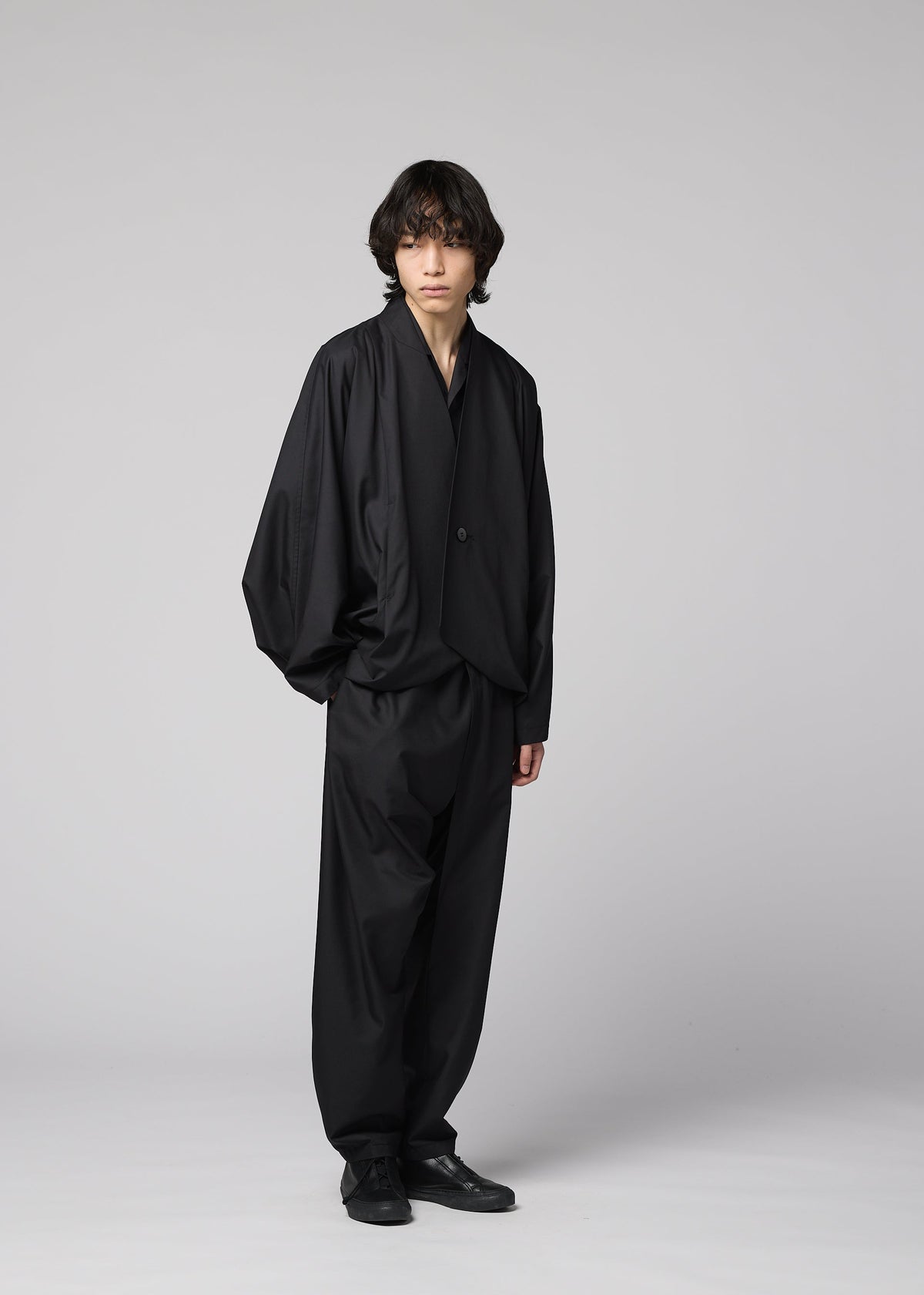 A person with medium-length dark hair stands against a light gray background, wearing IM MEN's SWING BLACK loose-fitting suit with wide, lightweight pants and black shoes. view 7 of 7