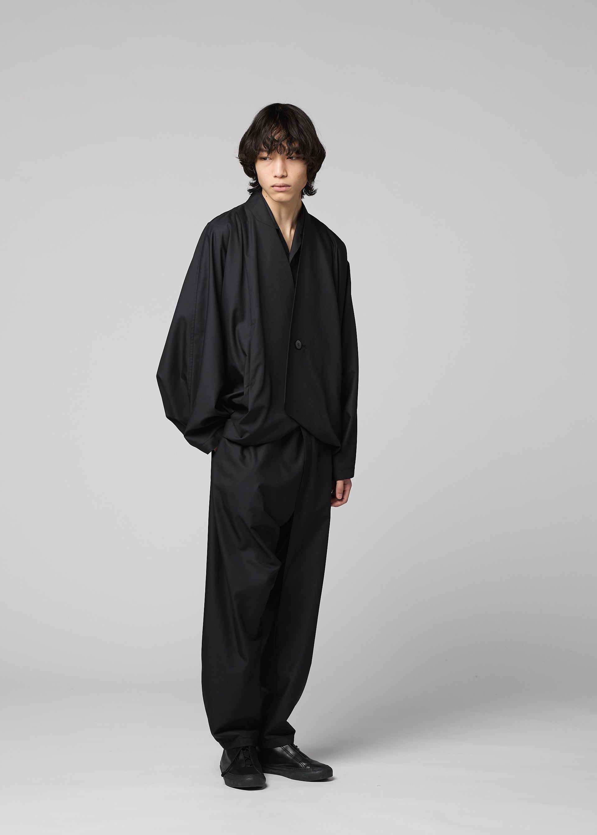 Zoomed view A person with medium-length dark hair stands against a light gray background, wearing IM MEN's SWING BLACK loose-fitting suit with wide, lightweight pants and black shoes.