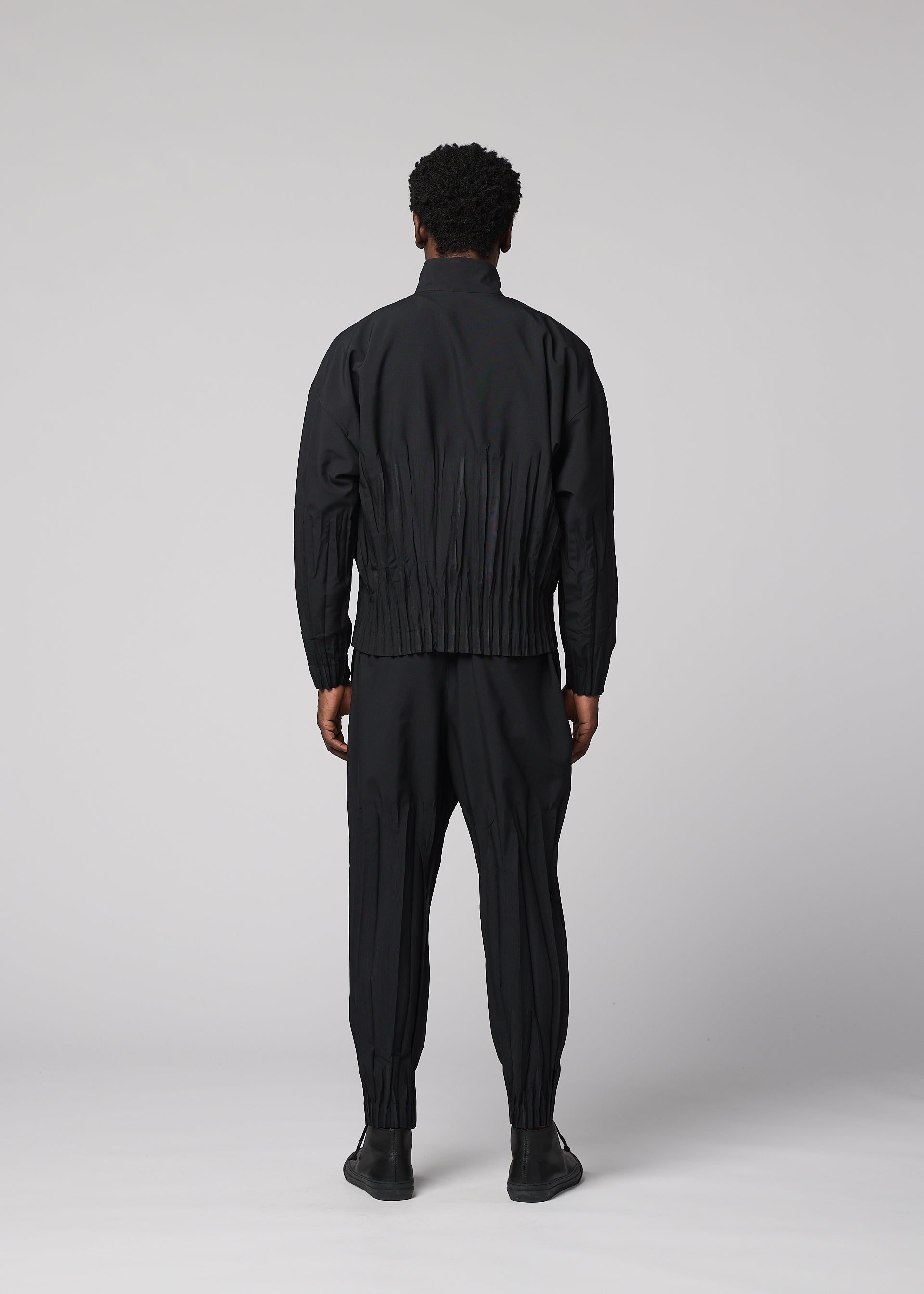 Zoomed view Facing away, a person with short curly hair wears the IM MEN WAFT BLACK set—loose-fitting jacket and matching black polyester pants with linen-like texture and gathered cuffs—paired with black shoes against a plain light gray background.