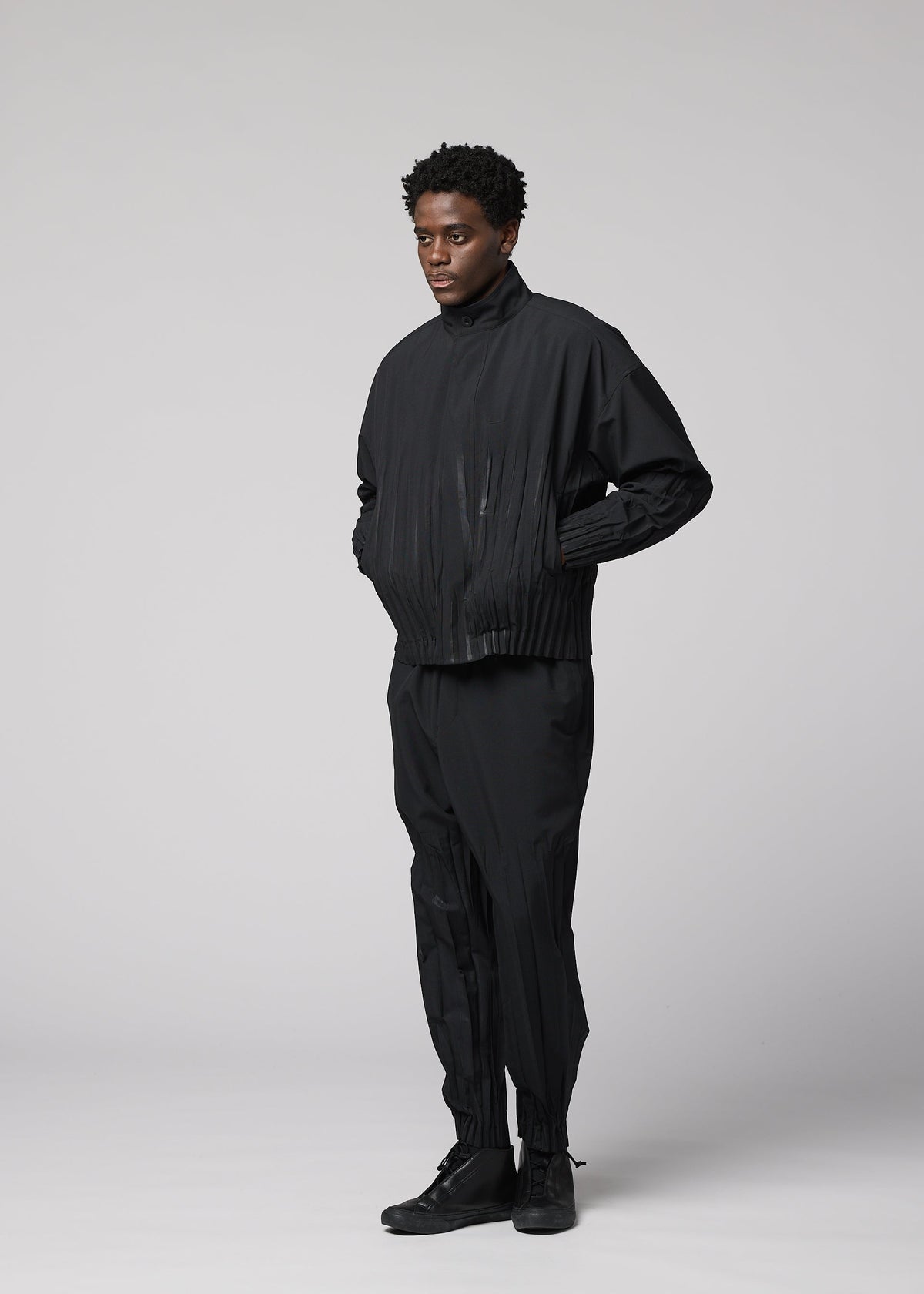 A person stands against a plain light gray background wearing IM MEN's WAFT BLACK: a loose-fitting black jacket and polyester pants with subtle pleats and linen-like texture, paired with black shoes. Hands in pockets, their expression is neutral. view 7 of 8