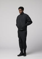 A person stands against a plain light gray background wearing IM MEN's WAFT BLACK: a loose-fitting black jacket and polyester pants with subtle pleats and linen-like texture, paired with black shoes. Hands in pockets, their expression is neutral.