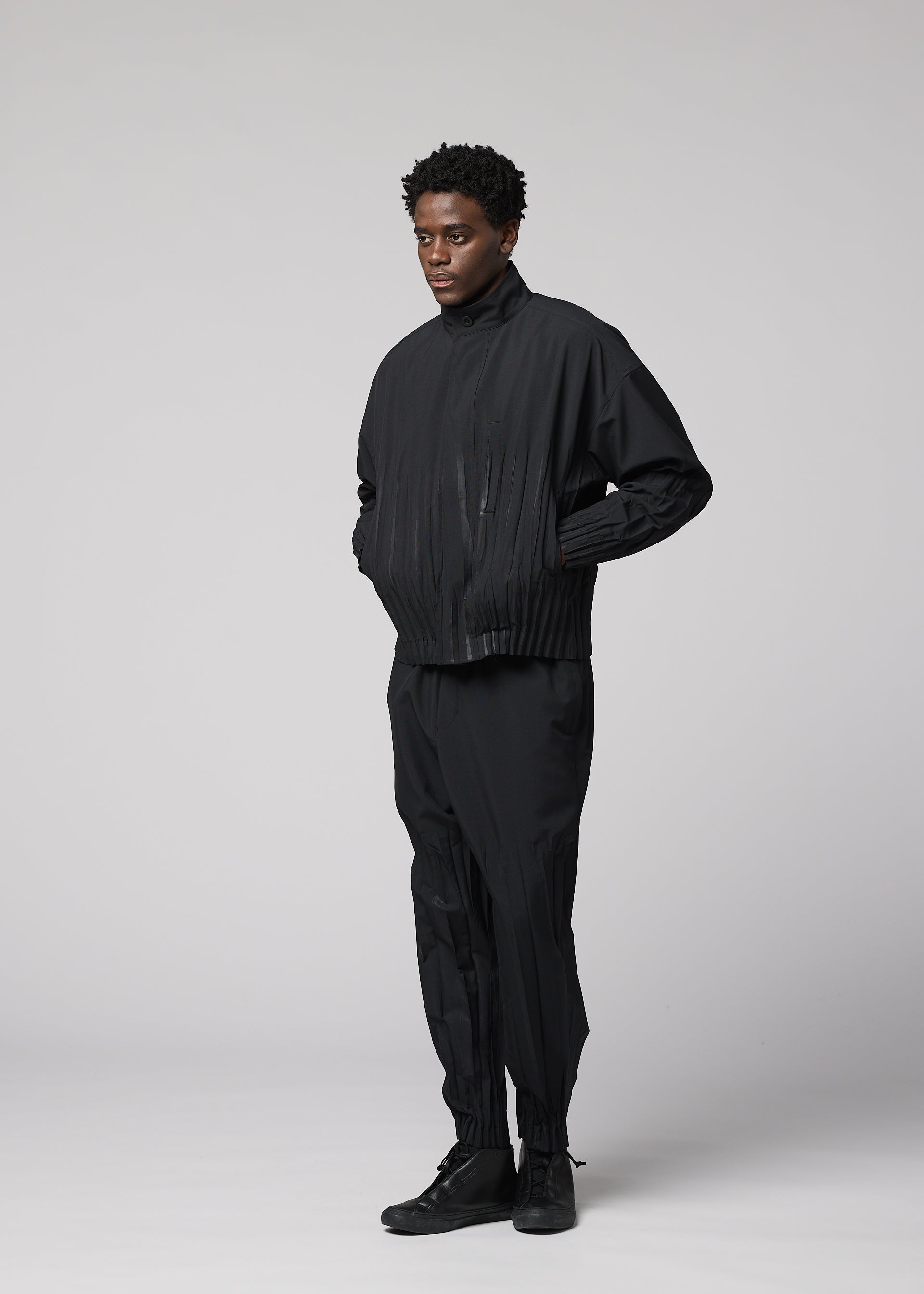 Zoomed view A person stands against a plain light gray background wearing IM MEN's WAFT BLACK: a loose-fitting black jacket and polyester pants with subtle pleats and linen-like texture, paired with black shoes. Hands in pockets, their expression is neutral.