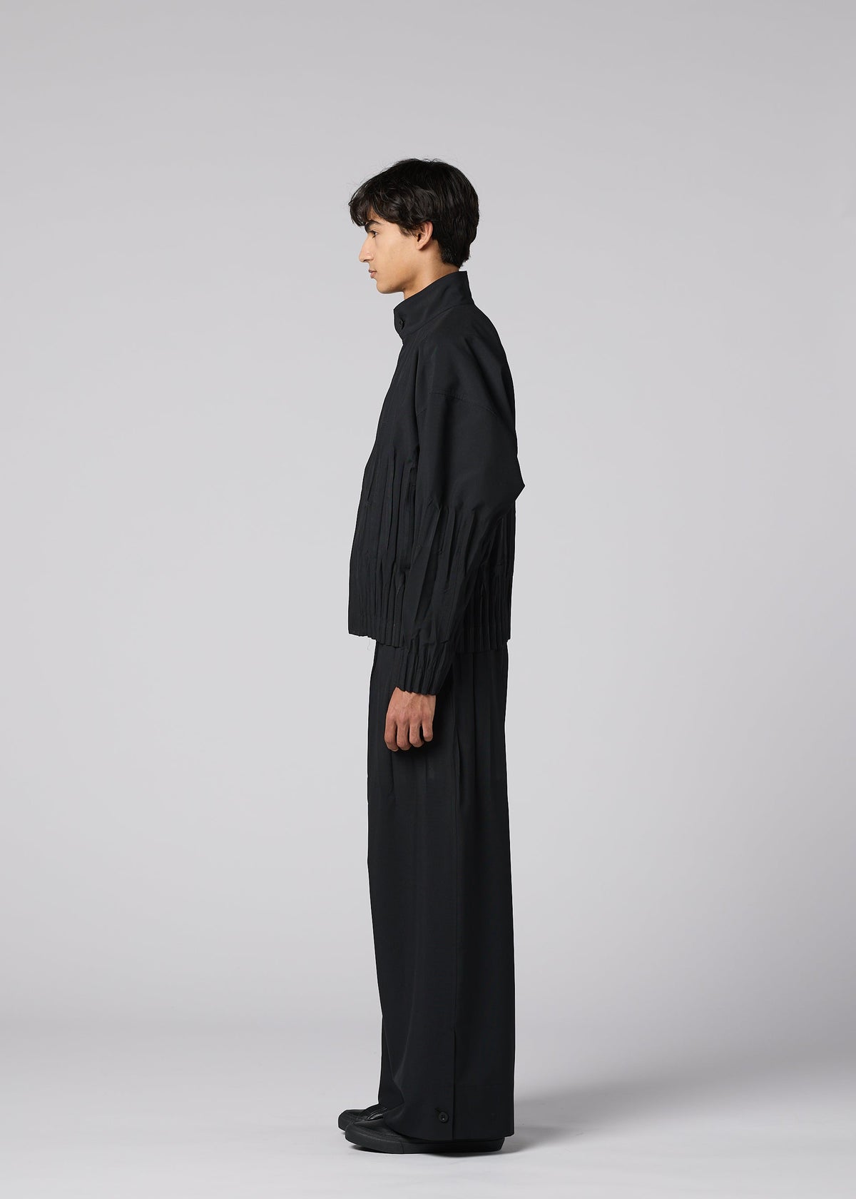 A person stands in profile against a light gray background, wearing IM MEN's WAFT BLACK: a loose-fitting black jacket and wide pants with adjustable hems. The minimalist outfit features a subtle linen-like texture for a chic, monochromatic look. view 5 of 9