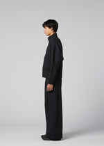 A person stands in profile against a light gray background, wearing IM MEN's WAFT BLACK: a loose-fitting black jacket and wide pants with adjustable hems. The minimalist outfit features a subtle linen-like texture for a chic, monochromatic look.