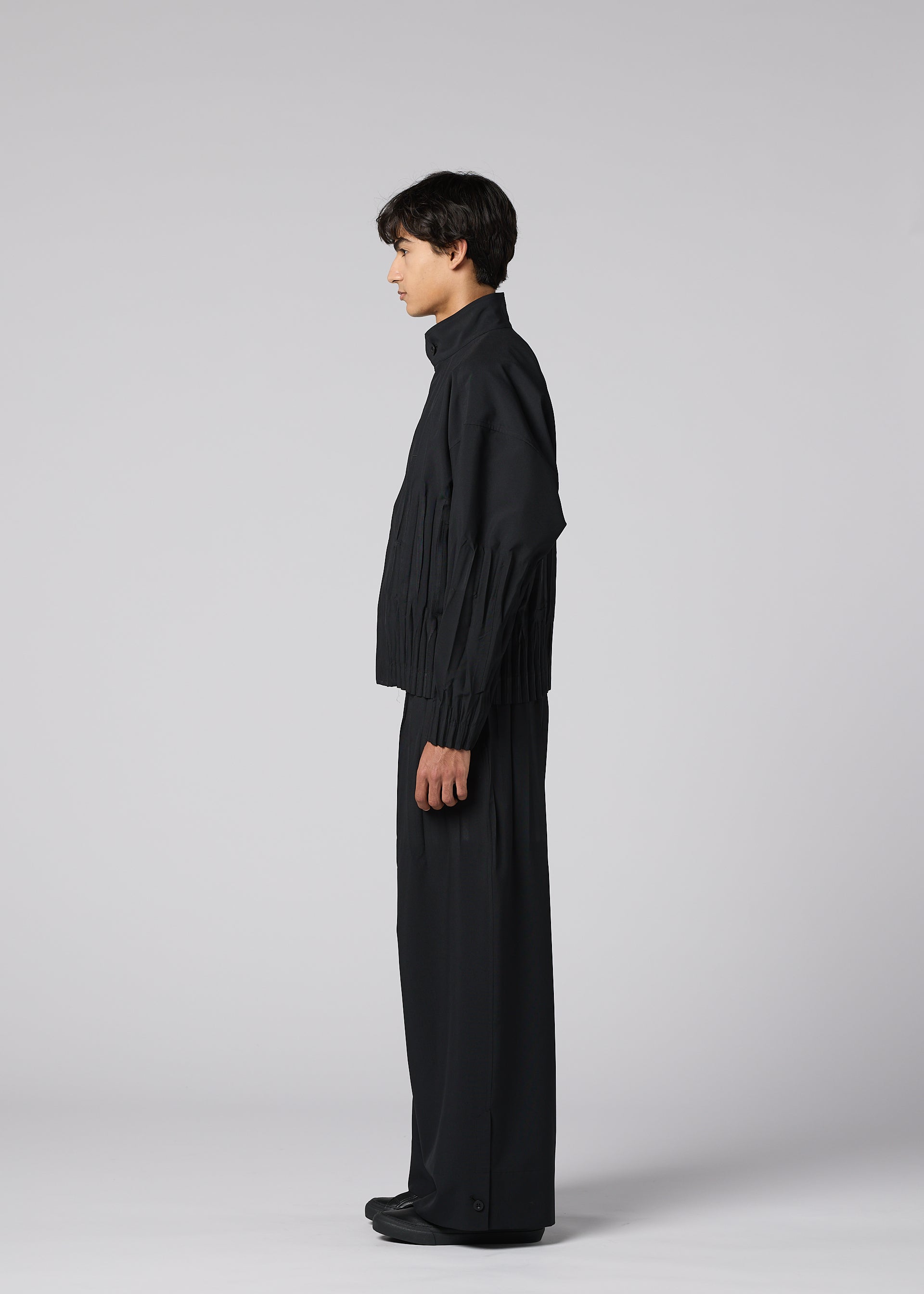 Zoomed view A person stands in profile against a light gray background, wearing IM MEN's WAFT BLACK: a loose-fitting black jacket and wide pants with adjustable hems. The minimalist outfit features a subtle linen-like texture for a chic, monochromatic look.