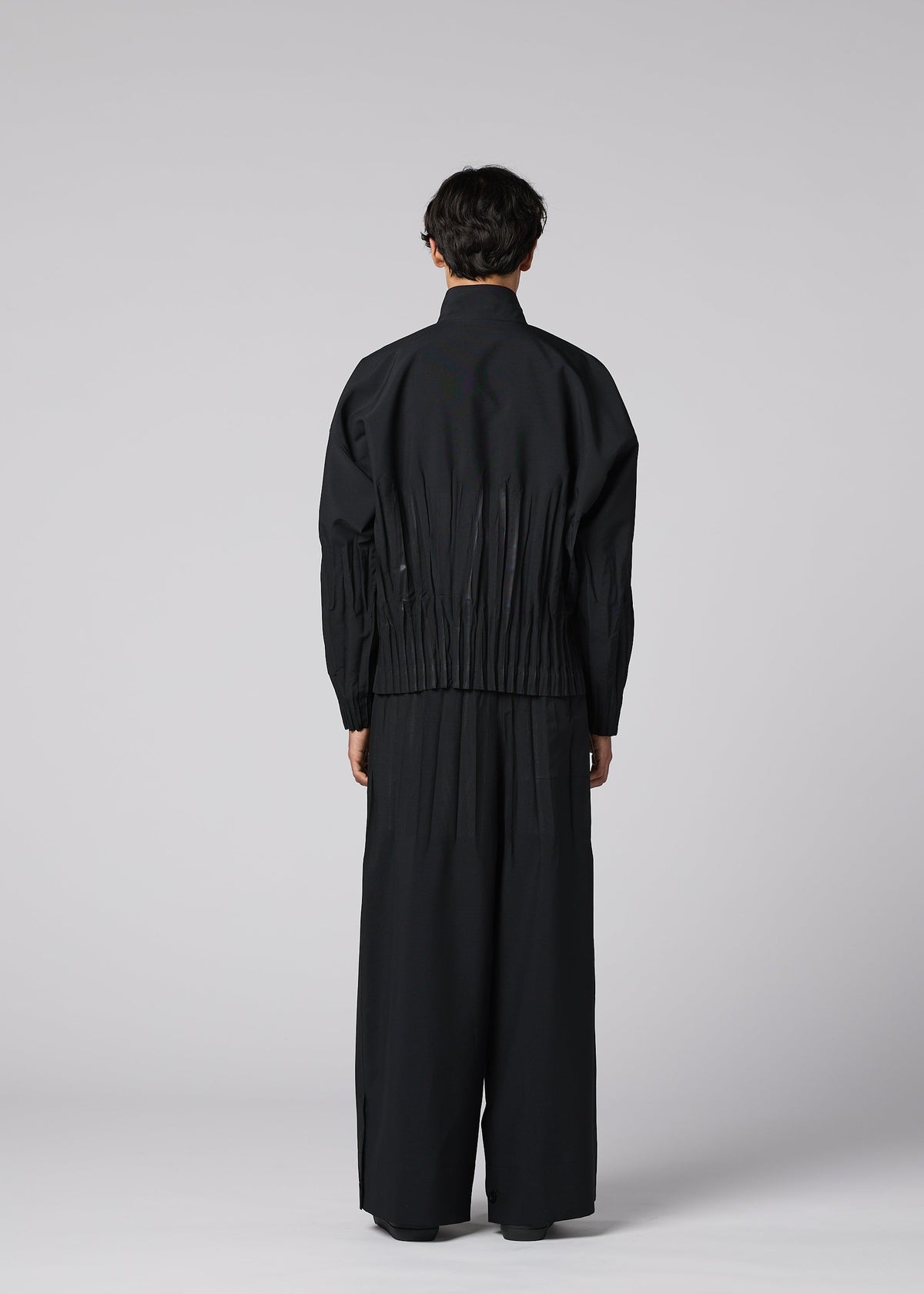 A person stands facing away, wearing IM MEN's WAFT BLACK— a loose-fitting black jacket with adjustable hem and gathered waist, paired with wide-leg, linen-textured black pants—set against a plain light gray background. view 6 of 9