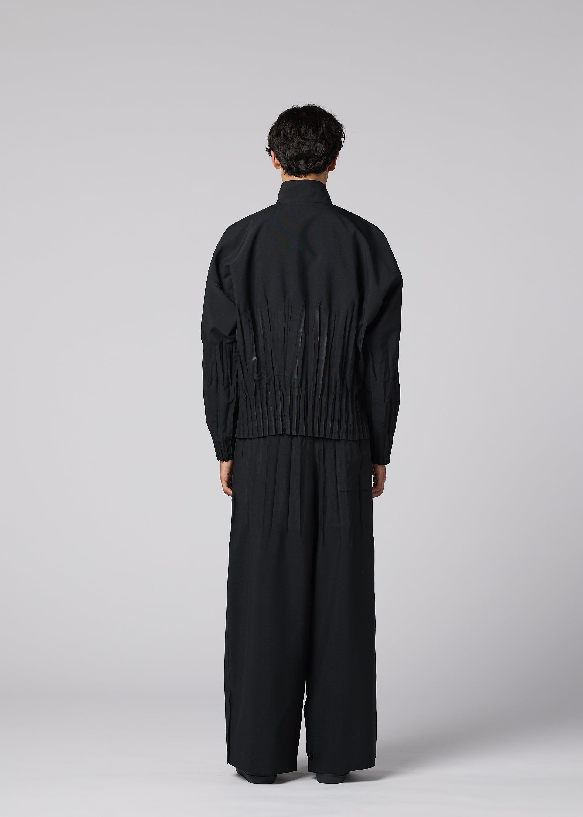 Zoomed view A person stands facing away, wearing IM MEN's WAFT BLACK— a loose-fitting black jacket with adjustable hem and gathered waist, paired with wide-leg, linen-textured black pants—set against a plain light gray background.