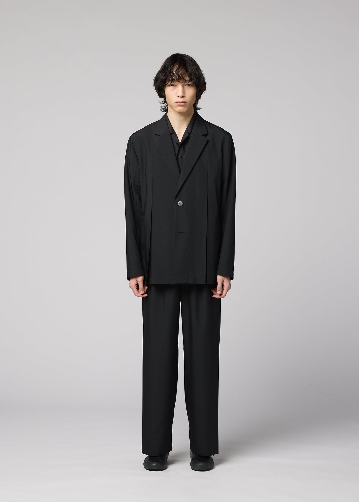 Zoomed view A person with medium-length dark hair faces forward, wearing the IM MEN WINGS BLACK oversized suit with wide pants and black shoes, against a plain light gray background.