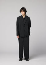 A person with medium-length dark hair stands against a gray background, wearing IM MEN’s WINGS BLACK wide, wrinkle-resistant pants and a loose-fitting black suit, one hand in their pocket and a neutral expression.