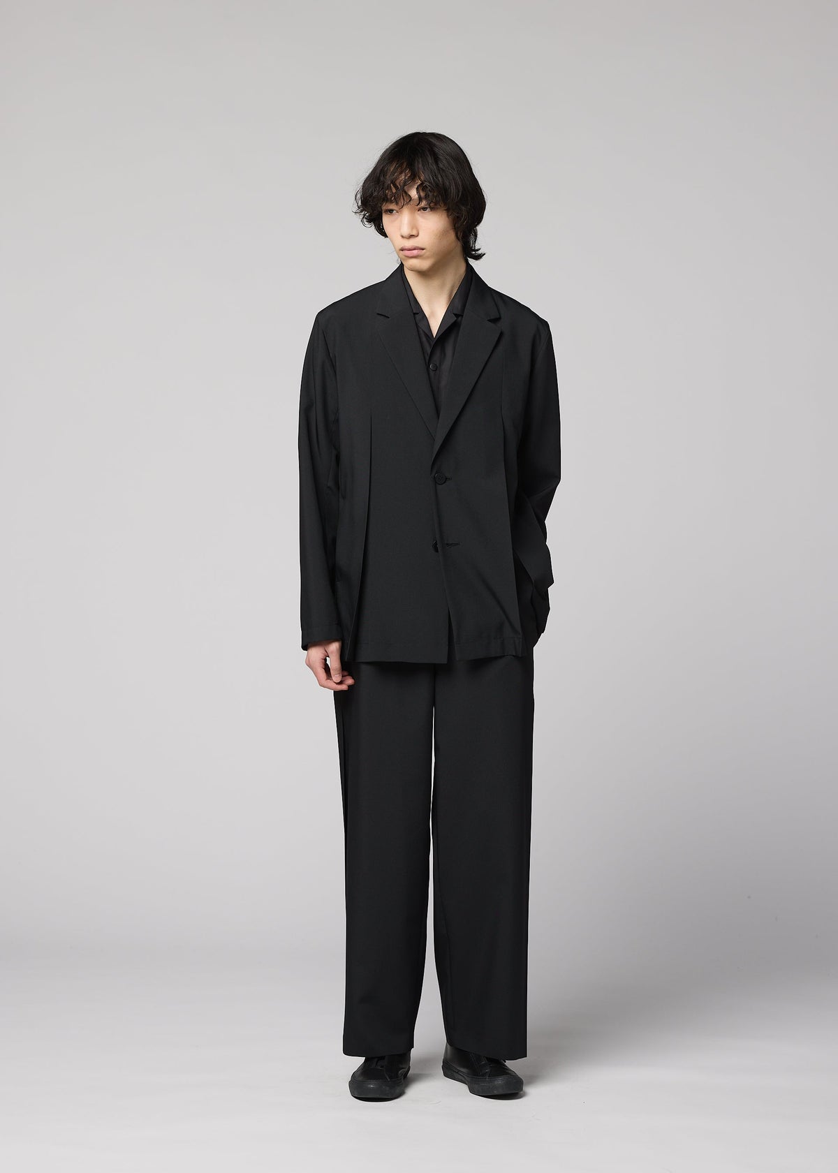 Zoomed view A person with medium-length dark hair stands against a gray background, wearing IM MEN’s WINGS BLACK wide, wrinkle-resistant pants and a loose-fitting black suit, one hand in their pocket and a neutral expression.
