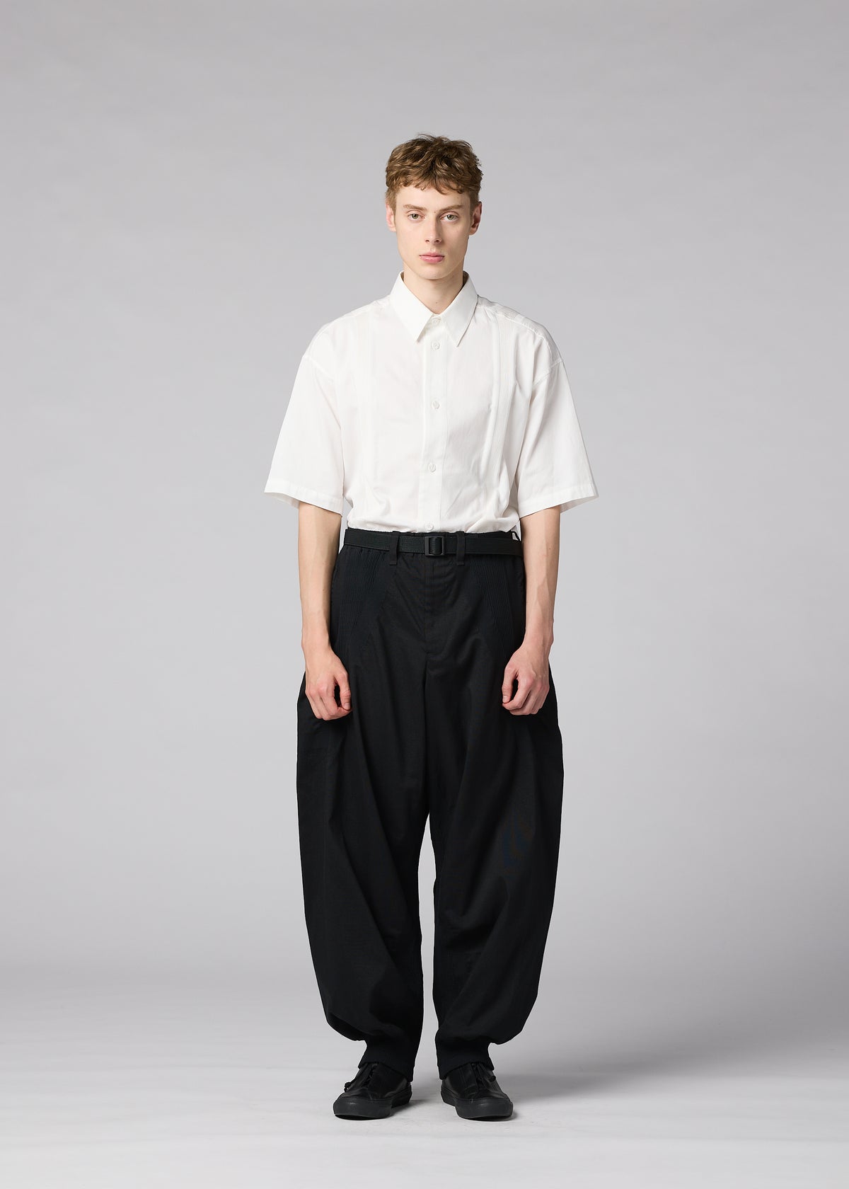 A person stands against a plain gray background, wearing the CLAY white short-sleeve button-up shirt by IM MEN, tucked into voluminous black pants with a belt and black shoes. view 5 of 8