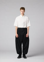 A person stands against a plain gray background, wearing the CLAY white short-sleeve button-up shirt by IM MEN, tucked into voluminous black pants with a belt and black shoes.