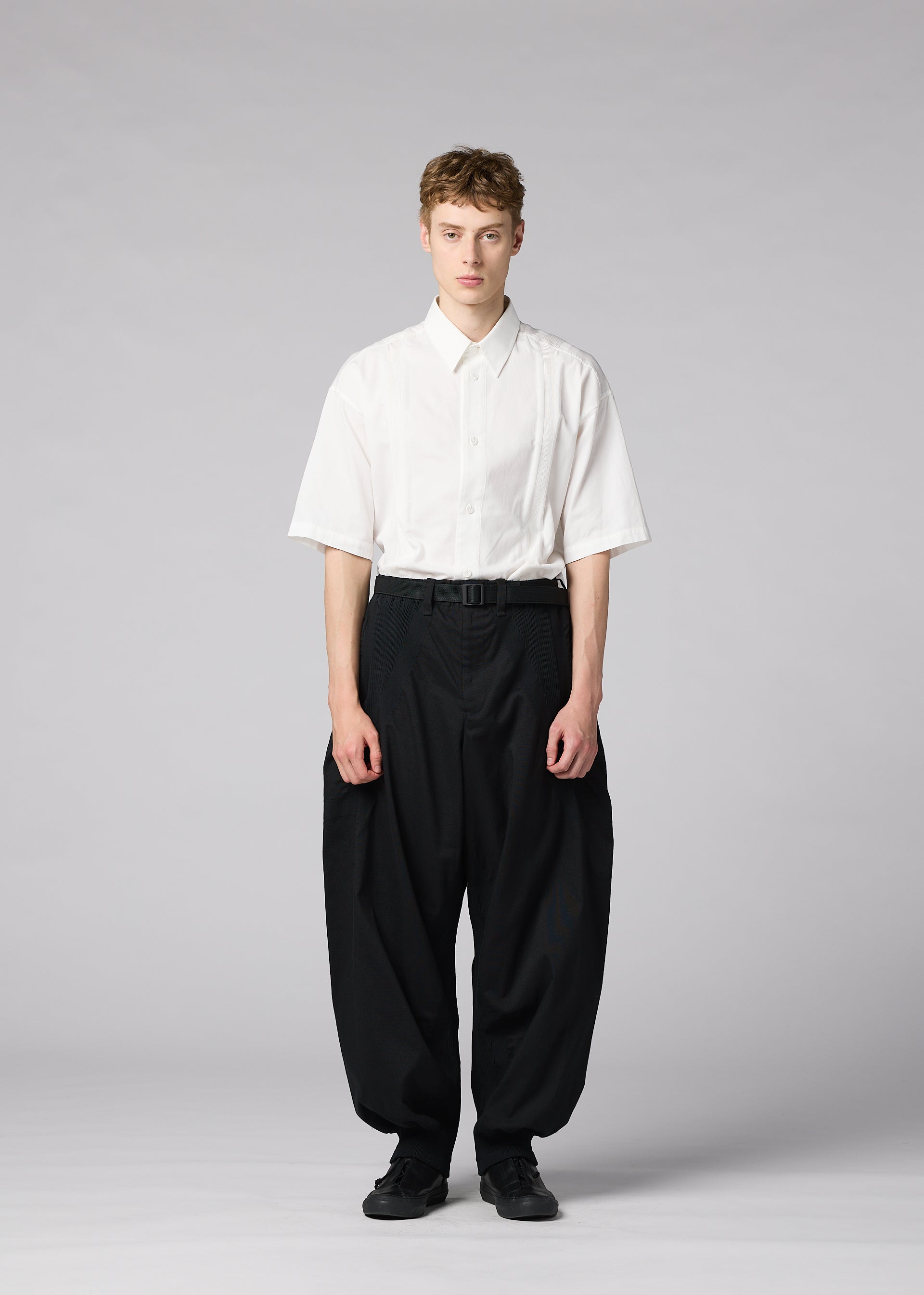 Zoomed view A person stands against a plain gray background, wearing the CLAY white short-sleeve button-up shirt by IM MEN, tucked into voluminous black pants with a belt and black shoes.