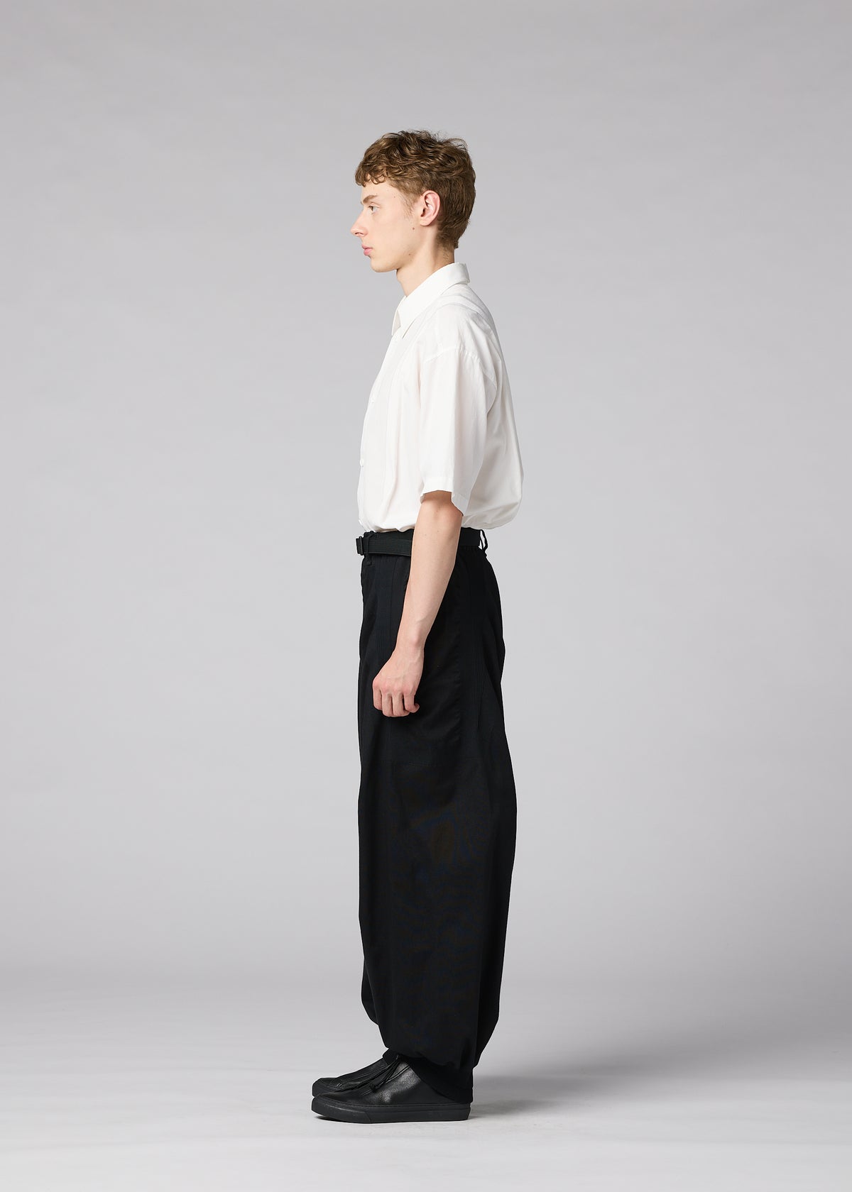 A young man stands in profile against a light gray background, wearing the IM MEN CLAY white short-sleeve shirt tucked into voluminous black pants and black shoes. view 6 of 8
