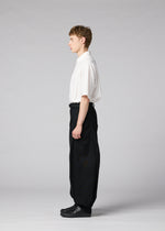 A young man stands in profile against a light gray background, wearing the IM MEN CLAY white short-sleeve shirt tucked into voluminous black pants and black shoes.