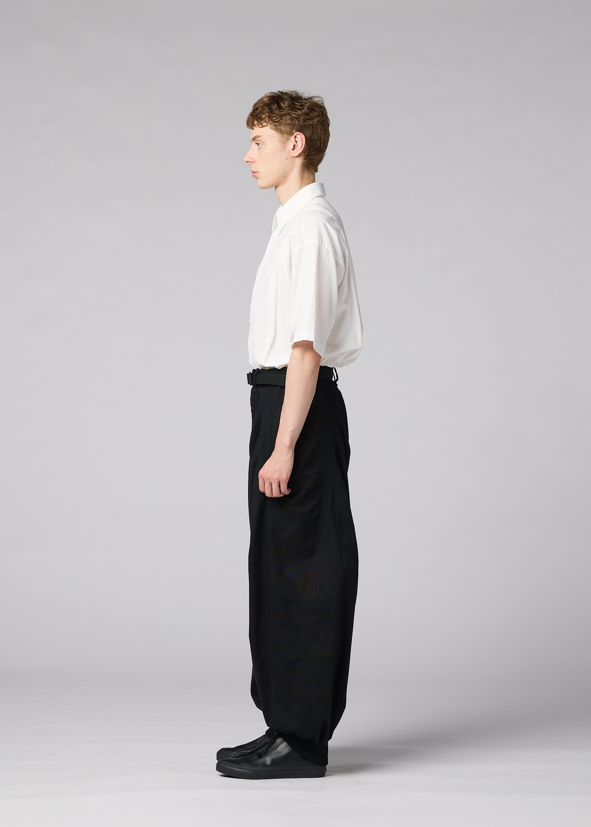 Zoomed view A young man stands in profile against a light gray background, wearing the IM MEN CLAY white short-sleeve shirt tucked into voluminous black pants and black shoes.