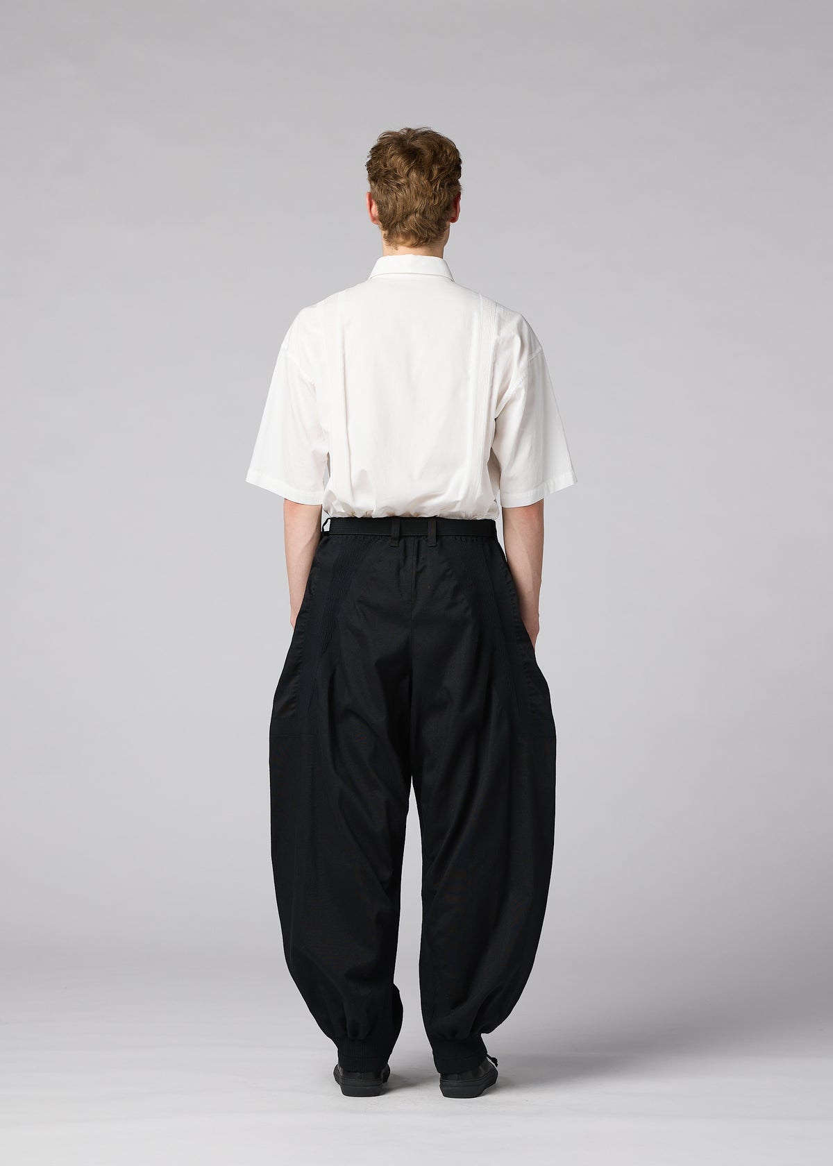A person stands facing away, dressed in IM MEN’s CLAY white short-sleeve shirt and loose, sculptural pants against a plain, light gray background. view 7 of 8