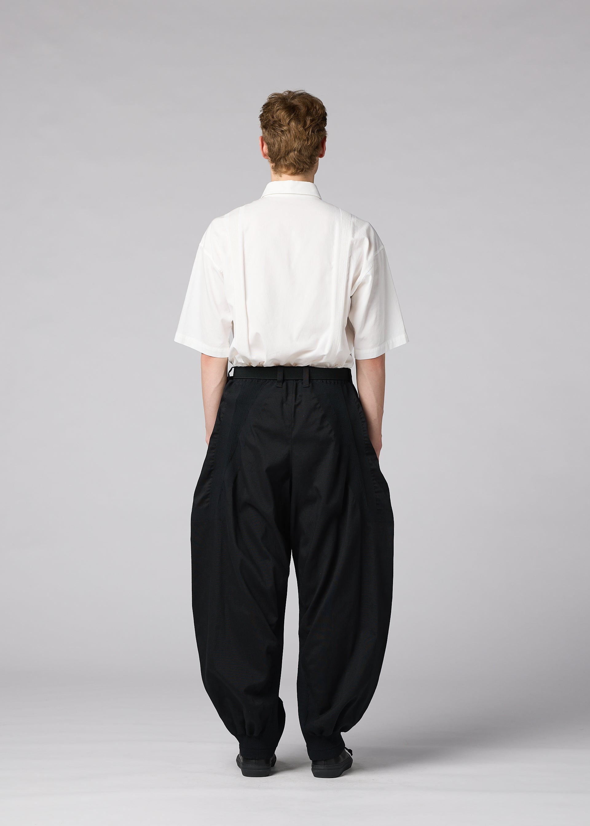Zoomed view A person stands facing away, dressed in IM MEN’s CLAY white short-sleeve shirt and loose, sculptural pants against a plain, light gray background.