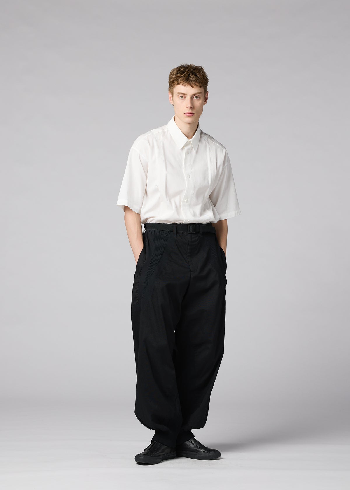 A person stands against a plain background, wearing IM MEN’s CLAY—a short-sleeved white button-up shirt tucked into voluminous black trousers with a belt, paired with black shoes. Their hands are in their pockets and their expression is neutral. view 8 of 8