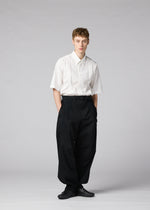 A person stands against a plain background, wearing IM MEN’s CLAY—a short-sleeved white button-up shirt tucked into voluminous black trousers with a belt, paired with black shoes. Their hands are in their pockets and their expression is neutral.