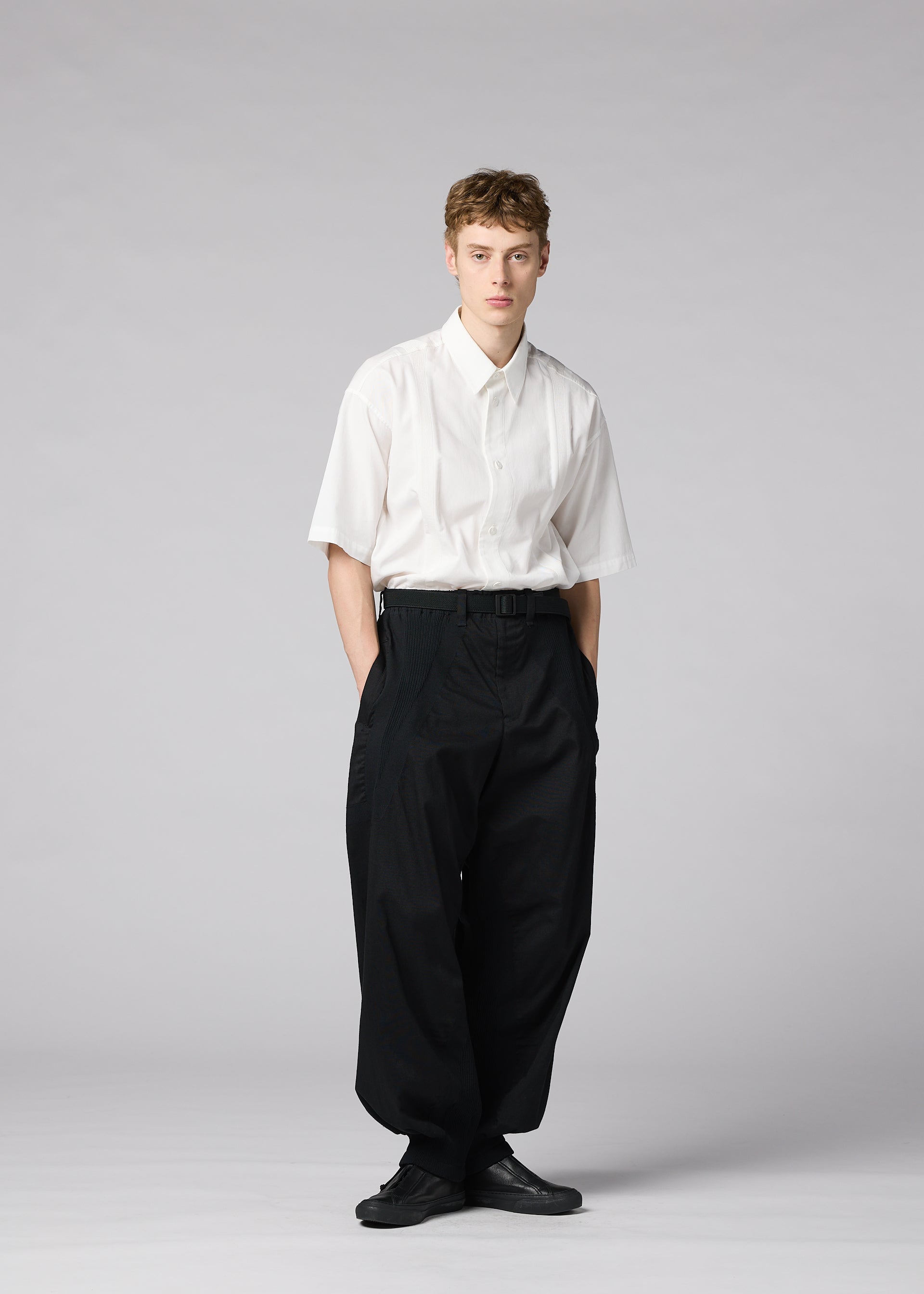 Zoomed view A person stands against a plain background, wearing IM MEN’s CLAY—a short-sleeved white button-up shirt tucked into voluminous black trousers with a belt, paired with black shoes. Their hands are in their pockets and their expression is neutral.
