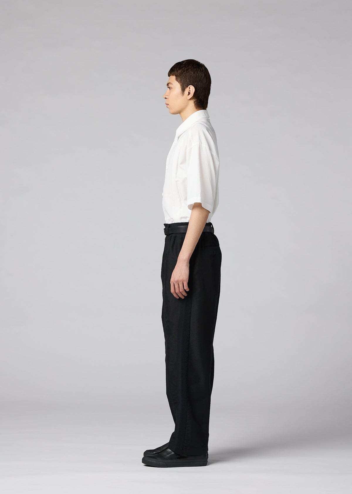 A person stands in profile facing left, wearing the IM MEN CLAY white short-sleeve button-up shirt, black tapered pants, and black shoes, against a plain gray background. view 6 of 8