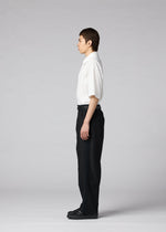 A person stands in profile facing left, wearing the IM MEN CLAY white short-sleeve button-up shirt, black tapered pants, and black shoes, against a plain gray background.
