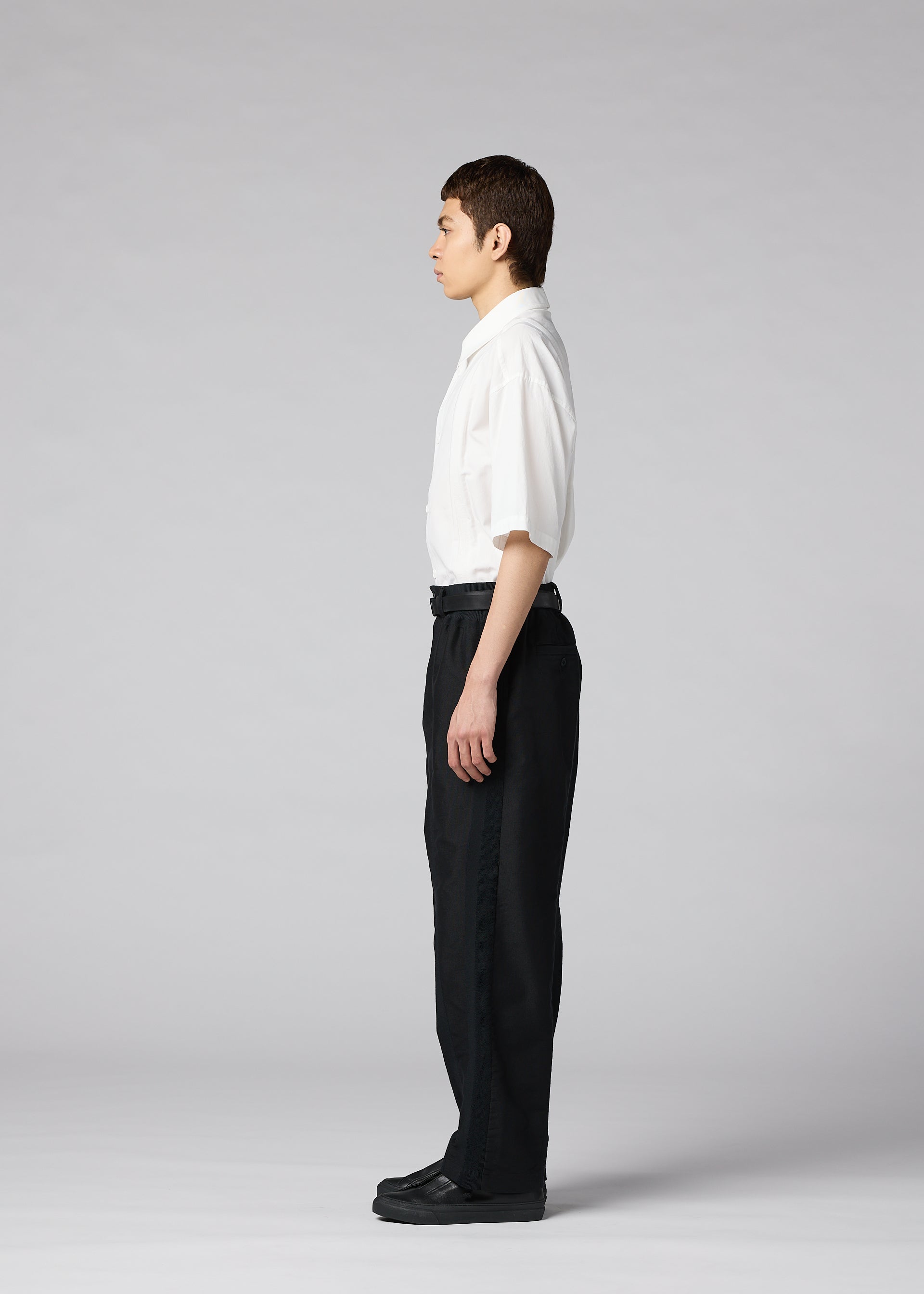Zoomed view A person stands in profile facing left, wearing the IM MEN CLAY white short-sleeve button-up shirt, black tapered pants, and black shoes, against a plain gray background.