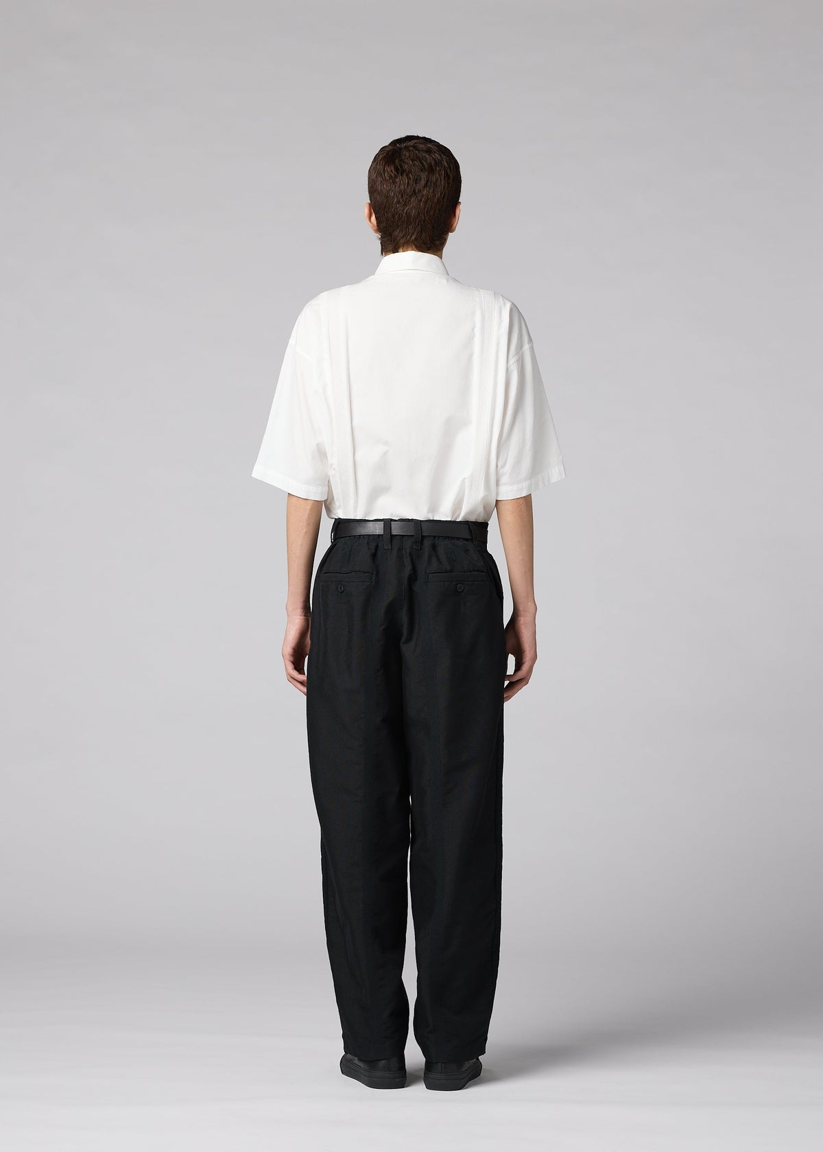 A person stands facing away on a plain light gray background, wearing the IM MEN “CLAY” loose white short-sleeve shirt tucked into high-waisted black tapered pants. view 7 of 8